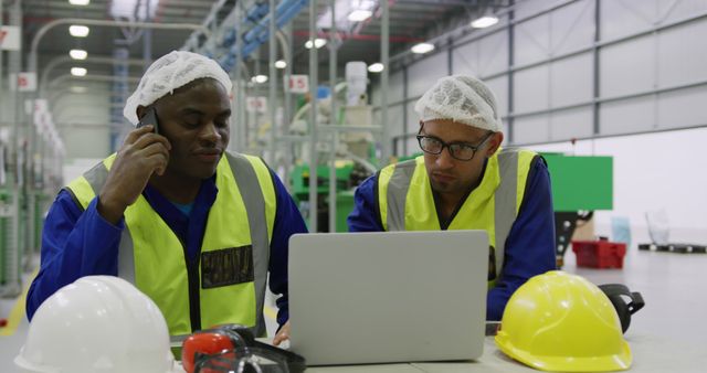 Two workers wearing safety vests and hairnets are collaborating in a modern warehouse, using a laptop for logistics and inventory management. This image is ideal for illustrating industrial teamwork, efficient warehouse operations, and the integration of technology in logistics. It can be used in articles, advertisements, or presentations related to manufacturing, logistics, supply chain management, and workplace safety.