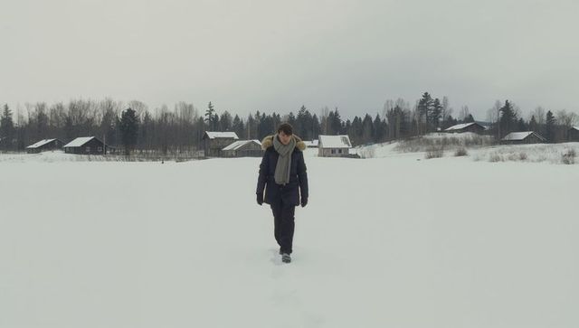 Man walking across snowy field toward camera leaving footprints near remote cabins, winter solitude