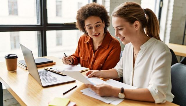 Two Women Collaborating Over Charts and Laptop in Modern Bright Office Workspace