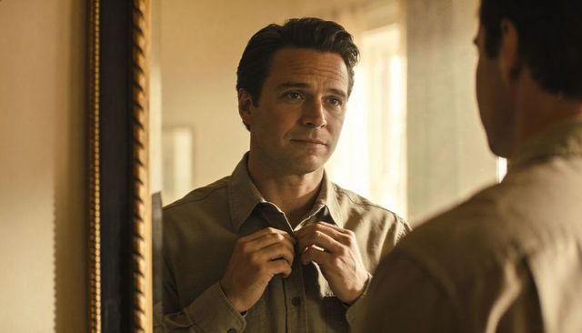Man buttoning shirt in mirror reflection, contemplative morning routine in neutral interior