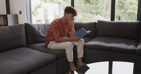 Young man reading notebook on modern sectional couch in bright contemporary living room