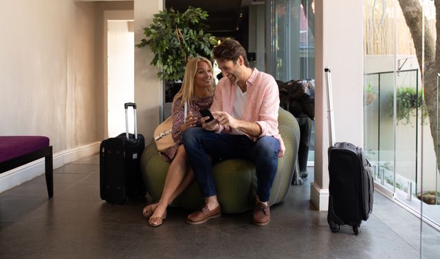 Front view of a Caucasian couple enjoying time off, arriving at a hotel on a sunny day, sitting together on a sofa in the lobby, the man using a smartphone, both smiling and talking