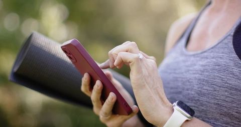 Mature woman holding yoga mat tapping smartphone wearing smartwatch outdoors