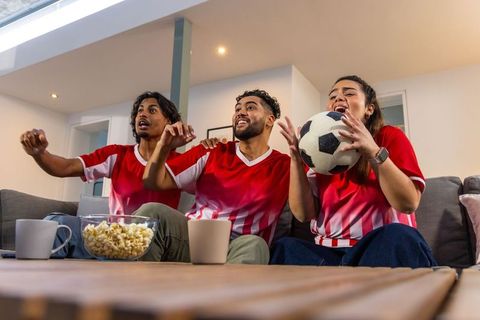 Friends cheering soccer match in living room wearing red jerseys holding ball and popcorn