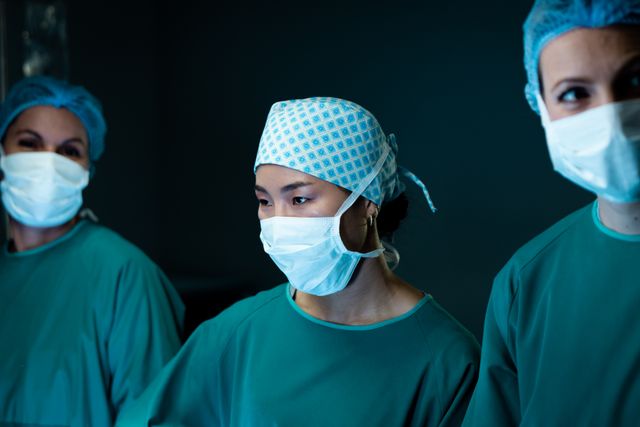 Three diverse female surgeons wearing face masks and caps, focused during a surgical procedure in an operating theatre. This image can be used for healthcare, medical services, hospital promotions, teamwork in healthcare, and patient care materials.
