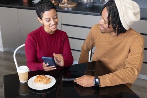 African american couple sharing tablet and smiling over coffee and croissant in cafe