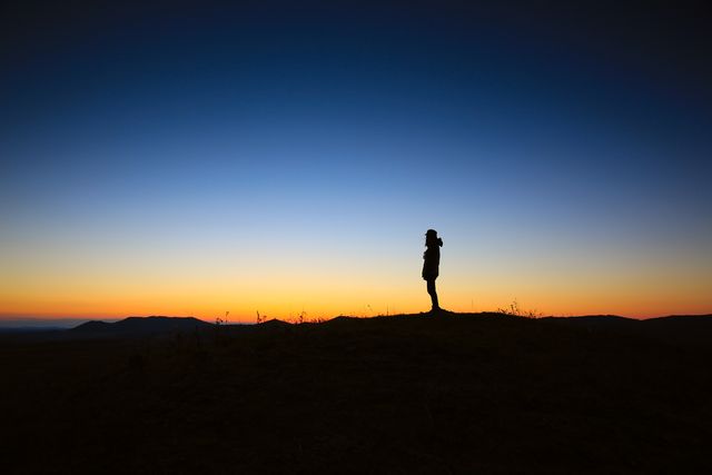 Person standing on mountain peak during sunset, creating a serene and contemplative atmosphere. Ideal for themes of solitude, nature, and tranquility. Great for advertisements, posters, blog posts, and motivational content.