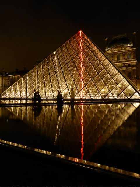 Louvre Pyramid illuminated with golden and red lights during nighttime with a stunning reflection in water. Ideal for travel advertisements, posters, architectural studies, or blogs about Parisian nightlife and iconic landmarks.