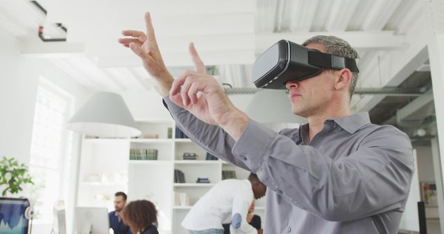 Senior businessman wearing virtual reality glasses in modern office, interacting with virtual objects using hand gestures. Modern work environment with colleagues working in the background. Ideal for concepts related to innovation in business, technology integration, or workplace dynamics.