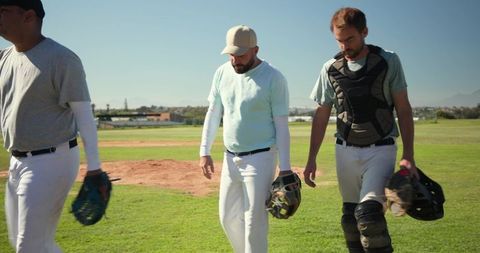 Baseball teammates walking off field holding gloves and catcher's gear in sunny league