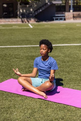 African american teen meditating cross-legged on pink yoga mat on green sports turf
