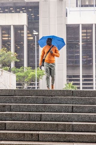 African american man walking steps in rain holding blue umbrella wearing orange sweater