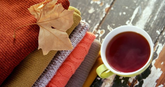 This image captures a cozy autumn scene with a stack of warm blankets, an autumn leaf, and a hot cup of tea on a rustic wooden table. Ideal for blog posts, social media, or websites focusing on seasonal themes, comfort, relaxation, or homely atmospheres.