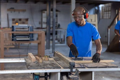 African American Carpenter Operating Table Saw Cutting Wood Planks with Safety Gear