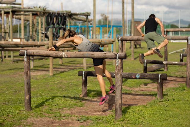 Rear view of a Caucasian woman and man wearing sports clothes vaulting over tall wooden hurdles at an outdoor gym during a bootcamp training session, with other outdoor gym equipment in the background