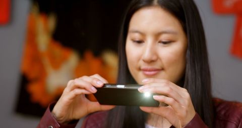 Image shows a young Asian woman focused on her smartphone while at home. Ideal for use in technology-related articles, online tutorials, lifestyle blogs focused on digital communication, or advertisements for mobile apps.