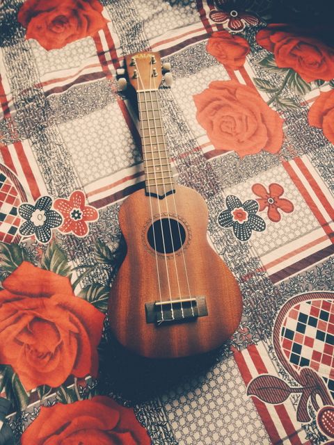 Ukulele resting on a vibrant, floral-patterned bedspread featuring red roses and various colorful patterns. This evokes a relaxed and cozy atmosphere. Ideal for promoting musical instruments, hobbies, home decor, or emphasizing relaxation and artistry in lifestyle contexts.