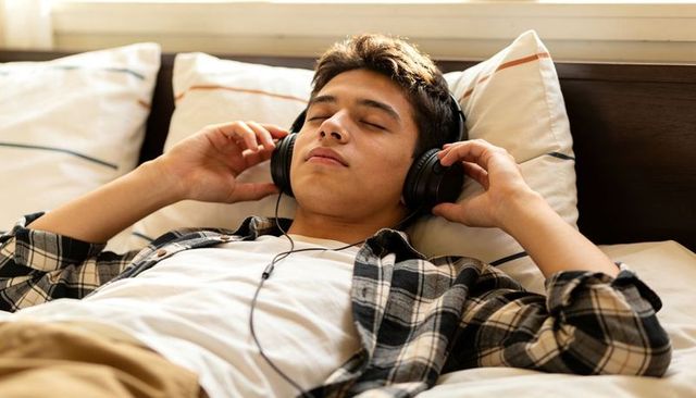 Male teen relaxing on bed listening to music with wired headphones, casual plaid shirt