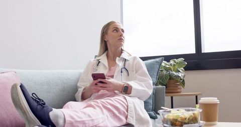 Nurse relaxing on lounge couch checking smartphone during staff break with coffee