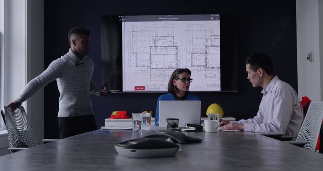 Three engineers are collaborating on a blueprint project in a modern, well-lit office. One is working on a laptop, another is using a tablet, and a third is presenting the blueprint plans displayed on a large screen mounted on the wall. This image is ideal for representing professional teamwork, modern office settings, architectural planning, and engineering discussions.