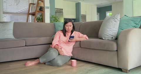 Mature woman sitting on floor beside sofa, scrolling smartphone with mug nearby