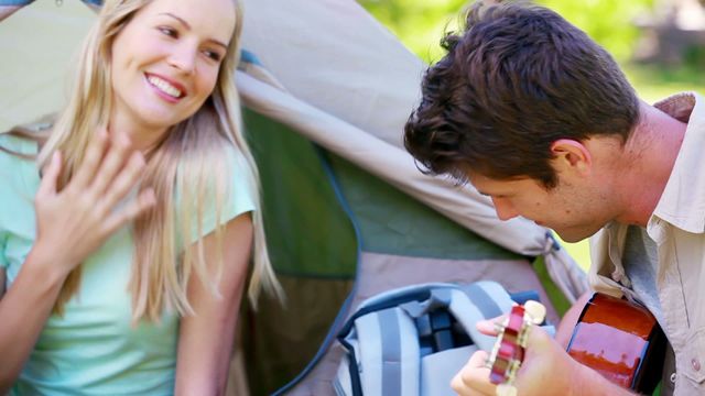 Couple sitting near tent, man playing guitar and singing, woman smiling. Perfect for promotions of outdoor activities, romantic getaways, music in nature, camping gear advertisements, and travel blogs highlighting couple's adventures.