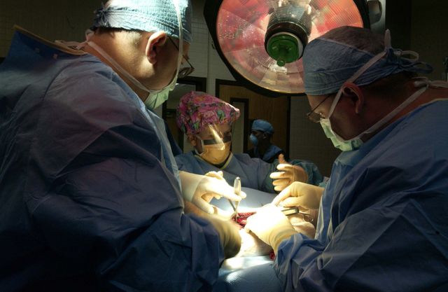Medical team performing surgery under strong operating light. Surgeons wearing masks, gloves, and protective clothing. Useful for healthcare articles, medical procedures information, and hospital promotional materials.
