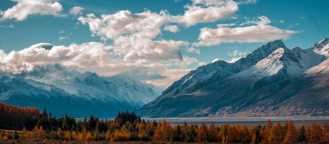 Beautiful autumn landscape featuring snow-capped mountain peaks and forested valley under partly cloudy sky. Ideal for use in travel brochures, nature blogs, outdoor adventure advertisements, and desktop wallpapers.