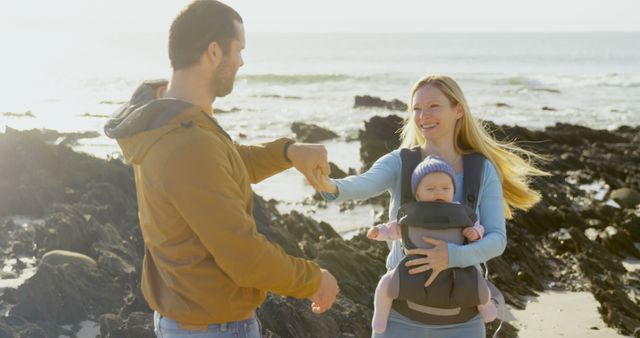 Happy caucasian father and mother with baby in baby carrier holding hands walking on sunny beach. Parenthood, family, care, wellbeing, nature, vacations and travel, unaltered.