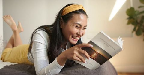Asian woman laughing while reading paperback, lying on bed in sunlit cozy bedroom