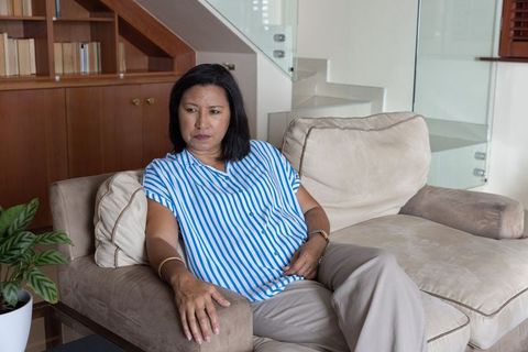 Mature woman sitting on beige sofa touching abdomen, pensive in modern cozy living room