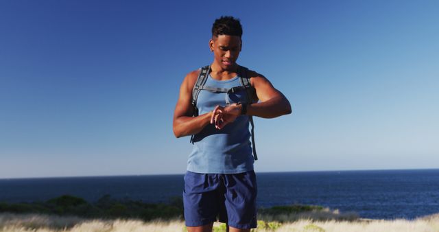 Young man in athletic wear with backpack is checking smartwatch on coastal hike, overlooking ocean. Ideal for fitness, outdoor activities, travel, and healthy lifestyle themes.