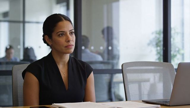 Hispanic businesswoman sitting in modern glass boardroom reviewing laptop and documents