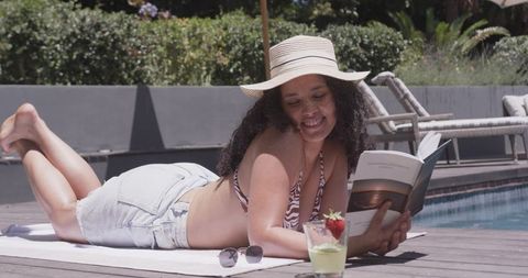 African american woman relaxing poolside reading paperback in sunhat and denim shorts
