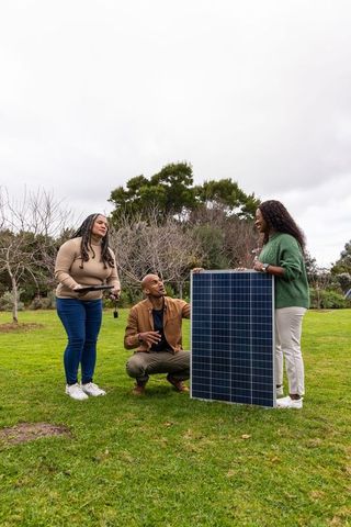African american technician inspecting large solar panel with homeowners on grassy lawn