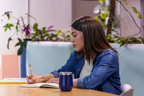 Asian woman writing in notebook at coworking space with plants, denim shirt, navy tumbler