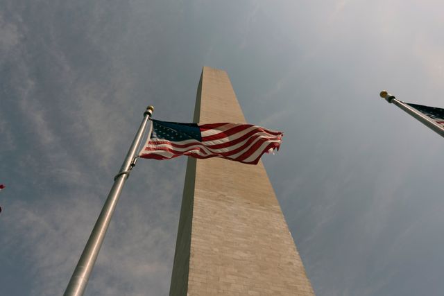 This photo depicts the Washington Monument standing tall with the US flag waving prominently in the foreground. It's captured from a low angle, giving a dramatic effect against a partly cloudy sky. Ideal for use in patriotic and nationalistic content, American history themes, tourists attractions promotions, and educational materials involving US landmarks.