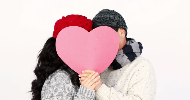 A couple, middle aged, is holding a large pink heart in front of their faces, with copy space. Their winter attire and the act of hiding behind the heart suggest a playful and romantic moment, related to Valentine's Day or love in general.