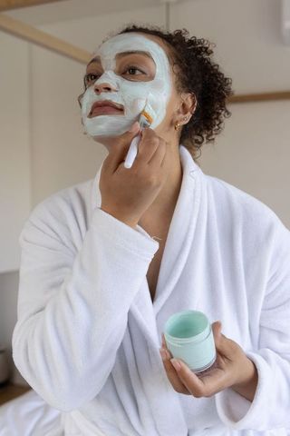 Woman applying mint facial mask with brush in robe for at-home spa selfcare routine