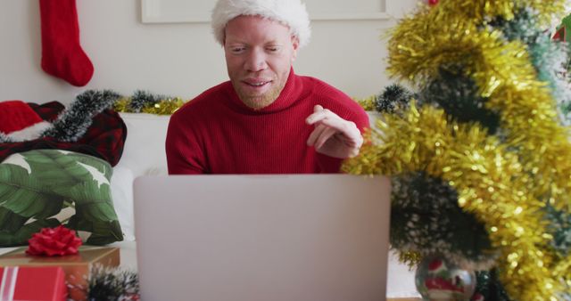 Man wearing Santa hat and red sweater having video call on laptop while surrounded by Christmas decorations. Perfect for themes related to holiday celebration, virtual social interactions, and festive gatherings.