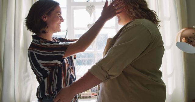Two women are enjoying a moment together as one fixes the other's shirt by a sunny window. This image is perfect for themes of friendship, care, and positive interactions. Suitable for use in blogs, social media posts, or advertisements promoting relationships and lifestyle content.
