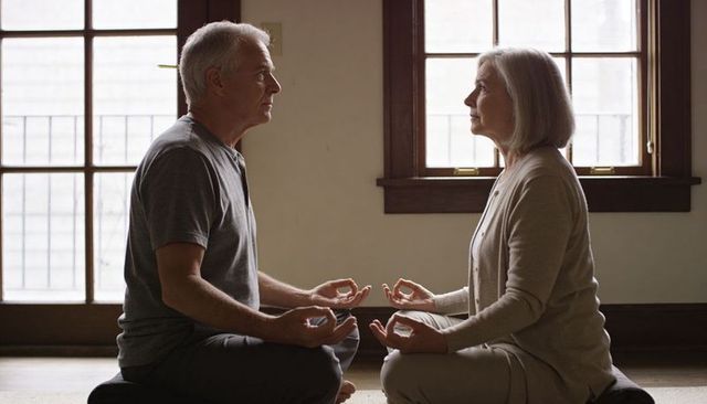 Older couple meditating together indoors on cushions by sunlit wooden windows for calm mindfulness