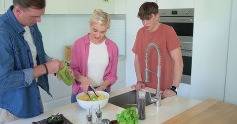Family cooking together at modern kitchen island mixing fresh pasta salad with teen son