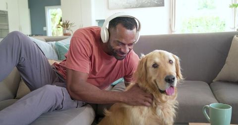 Mature african american man listening with headphones and petting golden retriever on sofa