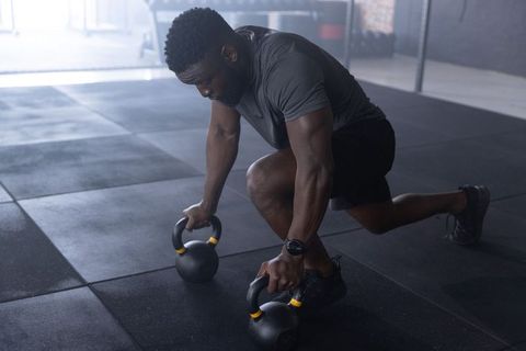 African american man performing kettlebell lunge wearing smartwatch on rubber gym floor