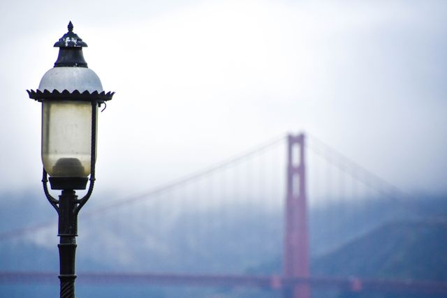 Vintage streetlamp on a foggy day with the Golden Gate Bridge in the background. Ideal for travel blogs, articles about San Francisco, posters, and tourism promotions highlighting famous landmarks and scenic locations in urban settings.
