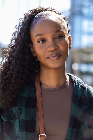 African woman gazing on city street wearing green-black plaid shirt and leather strap