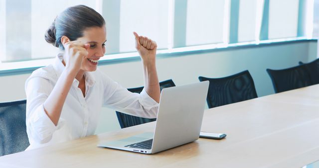 Depicts a businesswoman feeling excited and celebrating success while sitting at an office desk with open laptop. Ideal for promotions related to business achievements, employee motivation, corporate success stories, workplace productivity, and professional growth.