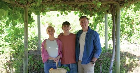 Family standing under vine pergola holding basket of fresh produce and garden tools