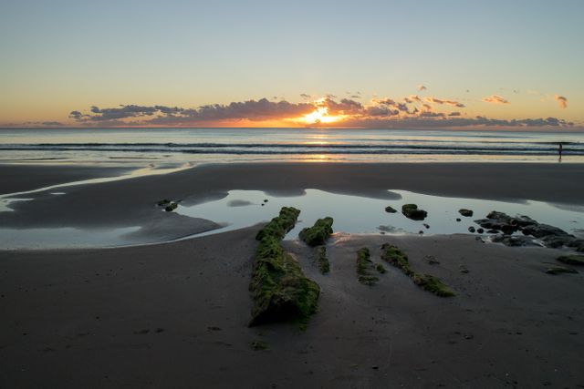 Sunrise over calm sea with reflections on wet sand creates a peaceful and serene scene. Low tide reveals rocks covered with algae. Ideal for travel adverts, relaxation themes, and nature photography.